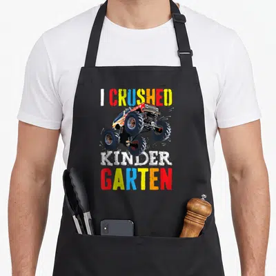 Graduation truck apron - I CRUSHED KINDER GARTEN text with cartoon monster truck in red yellow blue white — shown on a black cotton canvas apron against a white background
