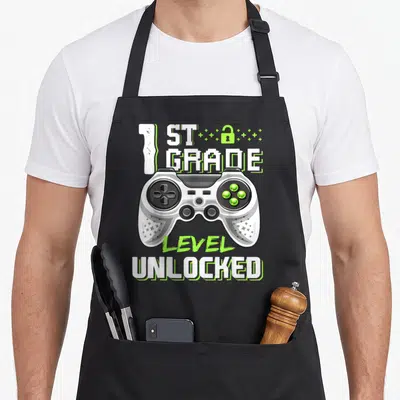 1st unlocked first apron - Game controller with 1st Grade Level Unlocked text and lock icon in white and lime green — shown on a black cotton canvas apron against a white background