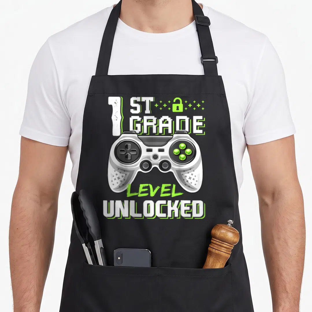 1st unlocked first apron - Game controller with 1st Grade Level Unlocked text and lock icon in white and lime green — shown on a black cotton canvas apron against a white background