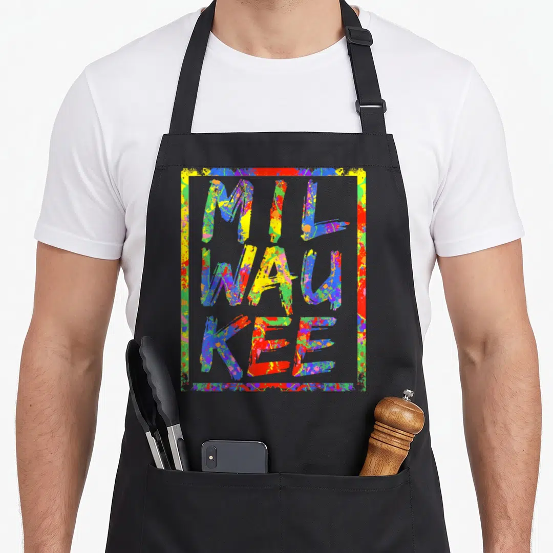 Paint milwaukee apron - Three stacked lines of text MIL WAU KEE filled with red yellow blue and green paint splatter — shown on a black cotton canvas apron against a white background