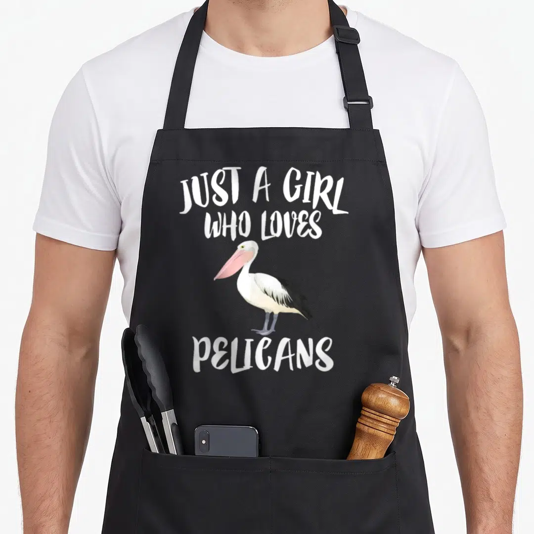 Funny pelican bird apron - Cartoon pelican with pink bill and text JUST A GIRL WHO LOVES PELICANS — shown on a black cotton canvas apron against a white background