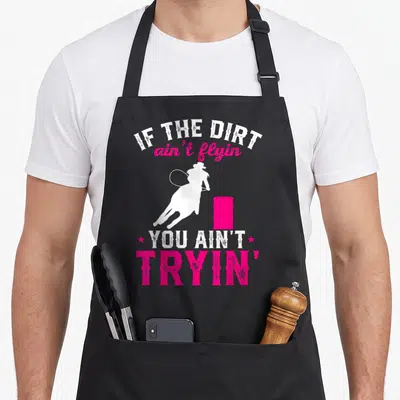 Barrel racing apron - White cowgirl silhouette on horse near pink barrel with distressed text IF THE DIRT ain't flyin — shown on a black cotton canvas apron against a white background