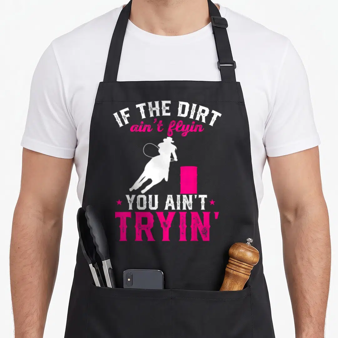 Barrel racing apron - White cowgirl silhouette on horse near pink barrel with distressed text IF THE DIRT ain't flyin — shown on a black cotton canvas apron against a white background