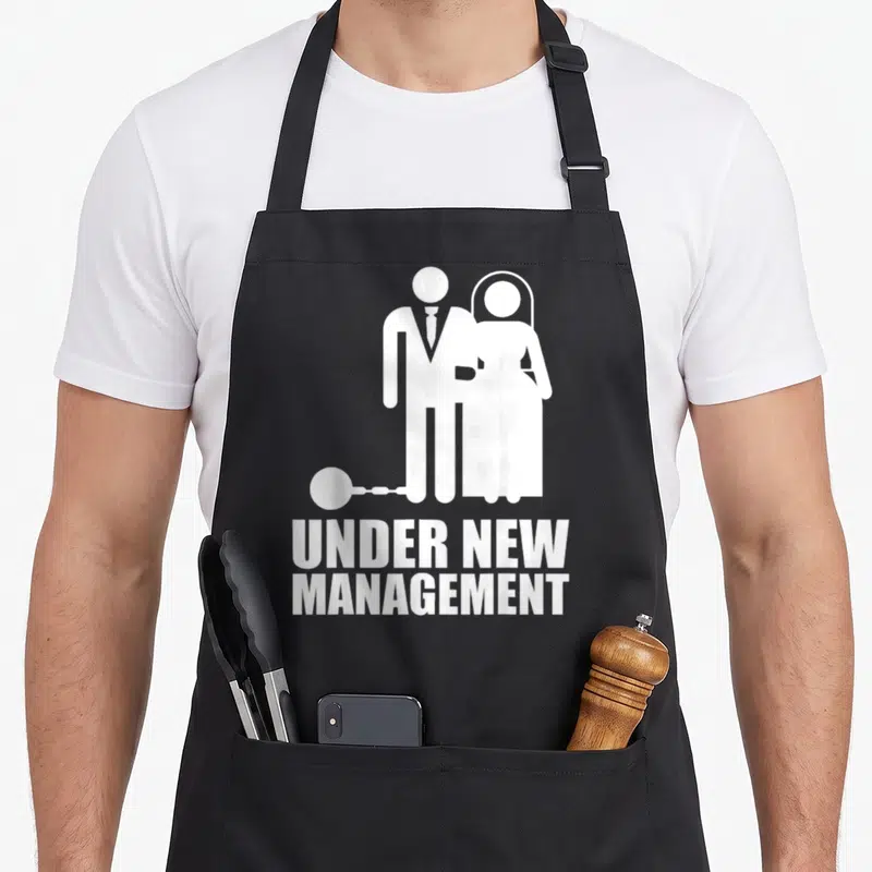 Married gag newlywed aprons - White couple silhouette with ball and chain and Under New Management text — shown on a black cotton canvas apron against a white background