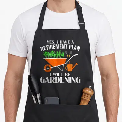 Gardening retirement apron - Orange wheelbarrow with green plants and Yes I Have A Retirement Plan text in distressed — shown on a black cotton canvas apron against a white background