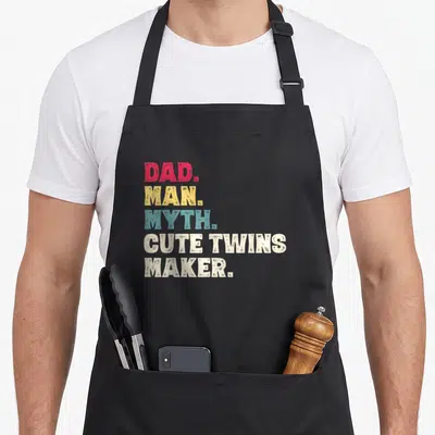 Myth makers apron - Stacked DAD MAN MYTH CUTE TWINS MAKER text in red yellow teal and white block letters — shown on a black cotton canvas apron against a white background