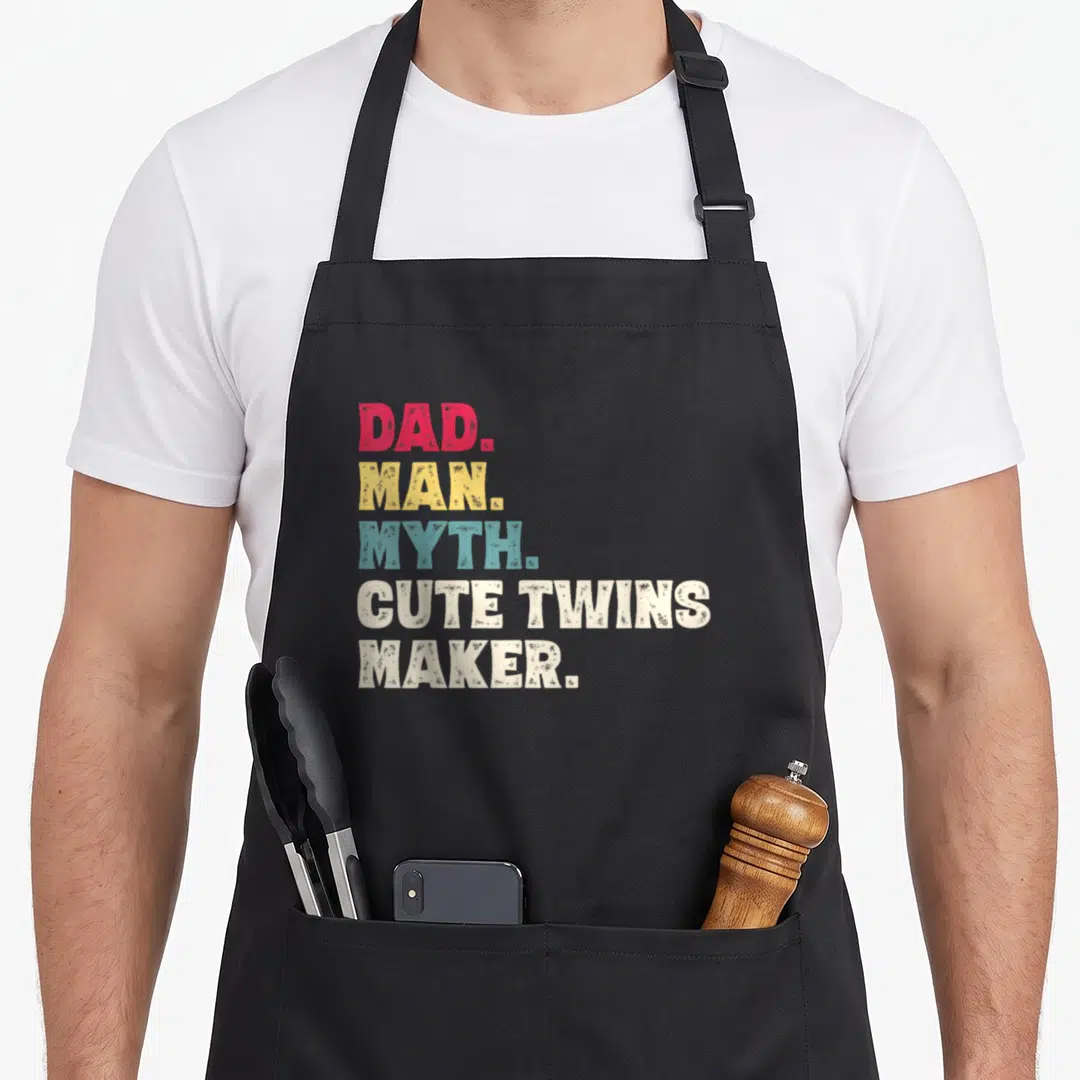 Myth makers apron - Stacked DAD MAN MYTH CUTE TWINS MAKER text in red yellow teal and white block letters — shown on a black cotton canvas apron against a white background