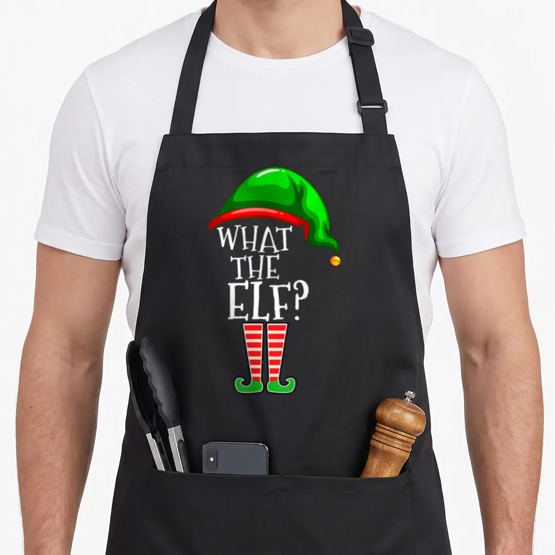 Family elf apron - What the elf text forming an elf body with green hat and striped legs — shown on a black cotton canvas apron against a white background