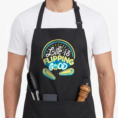 Arcade 80s apron - Stacked Life is Flipping Good text with two pinball flippers in yellow blue white cartoon art — shown on a black cotton canvas apron against a white background