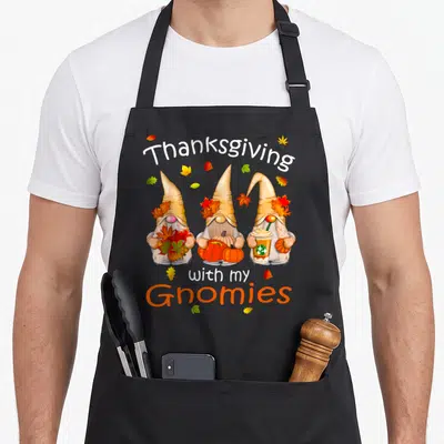 Gnome thanksgiving apron - Three gnomes holding pumpkins and leaves with text Thanksgiving with my Gnomies — shown on a black cotton canvas apron against a white background