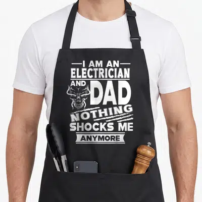 Shock electricians apron - White distressed text I Am An Electrician And Dad Nothing Shocks Me Anymore with skull — shown on a black cotton canvas apron against a white background