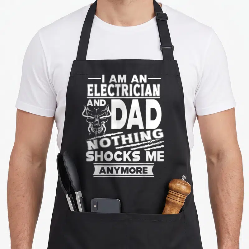 Shock electricians apron - White distressed text I Am An Electrician And Dad Nothing Shocks Me Anymore with skull — shown on a black cotton canvas apron against a white background