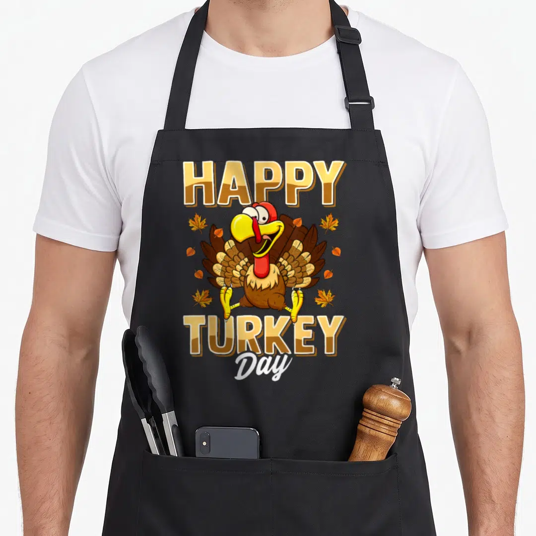 Turkey thanksgiving day aprons - Cartoon turkey surrounded by leaves with HAPPY TURKEY Day text in gold and brown — shown on a black cotton canvas apron against a white background