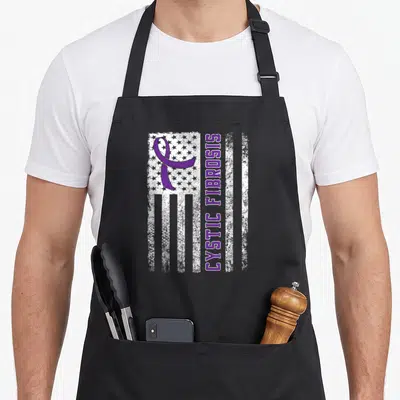 Cystic bow apron - Distressed white flag with purple ribbon and vertical CYSTIC FIBROSIS text — shown on a black cotton canvas apron against a white background