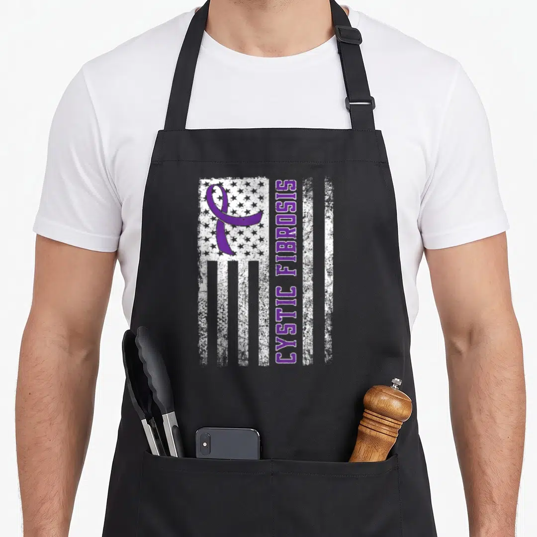 Cystic bow apron - Distressed white flag with purple ribbon and vertical CYSTIC FIBROSIS text — shown on a black cotton canvas apron against a white background