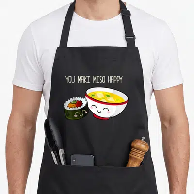 Maki kawaii apron - You Maki Miso Happy text with smiling sushi roll and miso soup bowl in kawaii art style — shown on a black cotton canvas apron against a white background