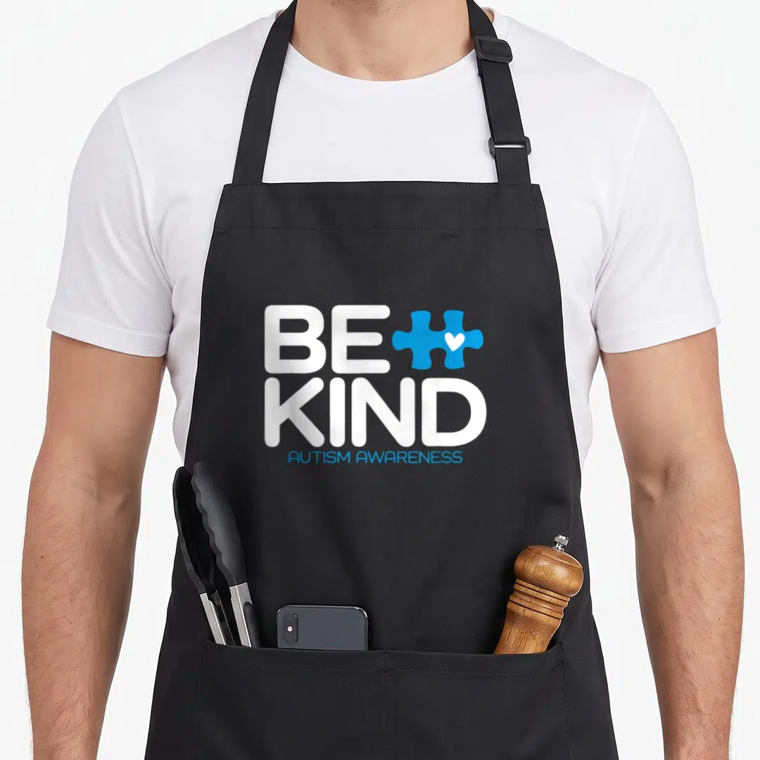 Kind autism awareness apron - Stacked white Be Kind text next to blue puzzle piece with white heart and Autism — shown on a black cotton canvas apron against a white background