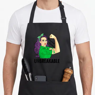 Warrior transplant awareness apron - Cartoon woman with purple hair and green ribbon tattoo, text Transplant Warrior — shown on a black cotton canvas apron against a white background