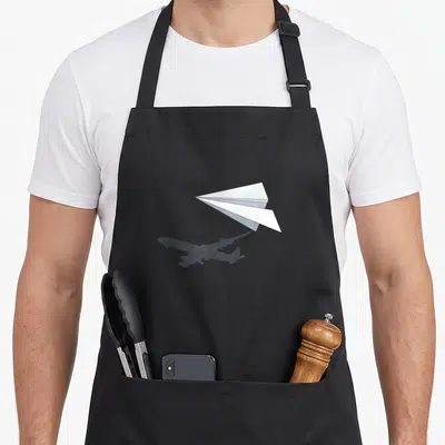 Controller airplane apron - White paper airplane above a dark grey commercial jet silhouette shadow — shown on a black cotton canvas apron against a white background
