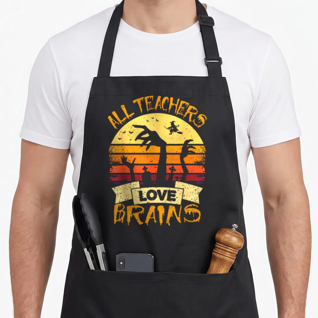 Brain teacher apron - Zombie hands rising over striped sunset with All Teachers Love Brains text and witch silhouette — shown on a black cotton canvas apron against a white background