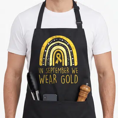 Childhood rainbow apron - Yellow white rainbow arch with gold ribbon and IN SEPTEMBER WE WEAR GOLD text — shown on a black cotton canvas apron against a white background