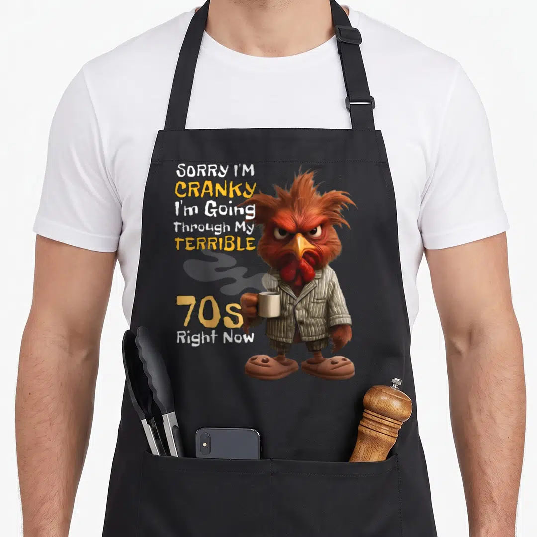 Through 70s apron - Stacked text sorry im cranky next to grumpy rooster in pajamas holding coffee mug — shown on a black cotton canvas apron against a white background
