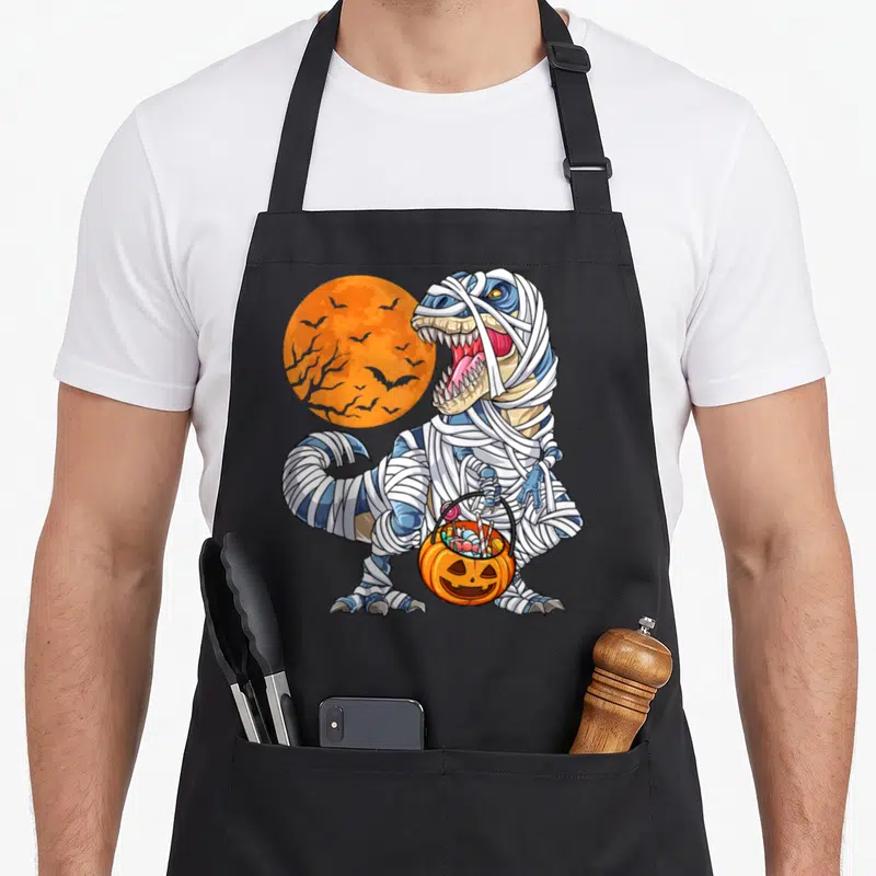 Halloween mummy and me aprons - Blue mummy t-rex holding pumpkin candy bucket in front of orange moon — shown on a black cotton canvas apron against a white background