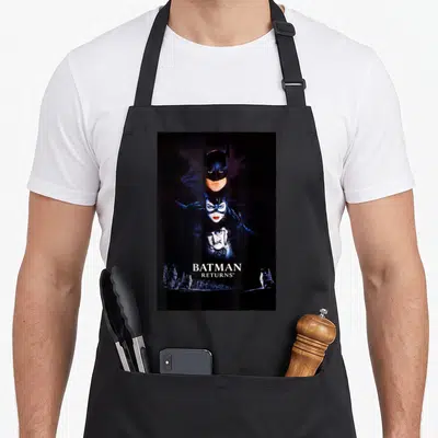 Return penguin aprons - Vertical stack of Batman, Catwoman, Penguin faces with title and penguin figures in black white — shown on a black cotton canvas apron against a white background