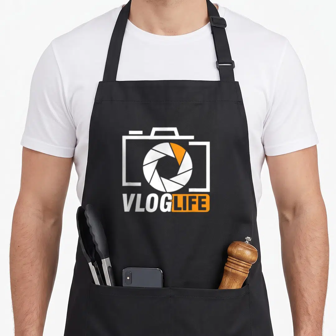 Influencer vlogging apron - White camera icon and VLOGLIFE text with orange rectangle accent on black fabric — shown on a black cotton canvas apron against a white background