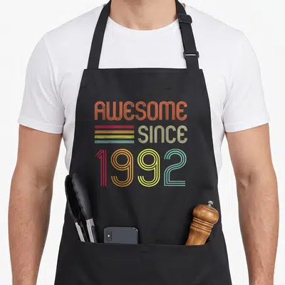 31st birthday apron - Retro typography reading AWESOME SINCE 1992 in orange beige red yellow green and blue lines — shown on a black cotton canvas apron against a white background
