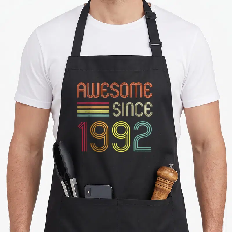 31st birthday apron - Retro typography reading AWESOME SINCE 1992 in orange beige red yellow green and blue lines — shown on a black cotton canvas apron against a white background
