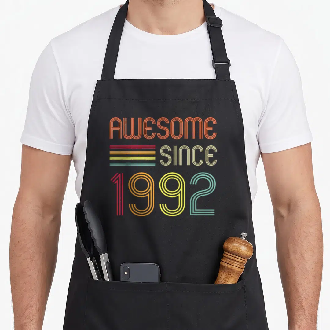 31st birthday apron - Retro typography reading AWESOME SINCE 1992 in orange beige red yellow green and blue lines — shown on a black cotton canvas apron against a white background