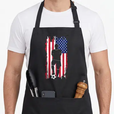 Soccer american flag apron - Grey soccer player silhouette standing on a ball against a distressed red white and blue — shown on a black cotton canvas apron against a white background