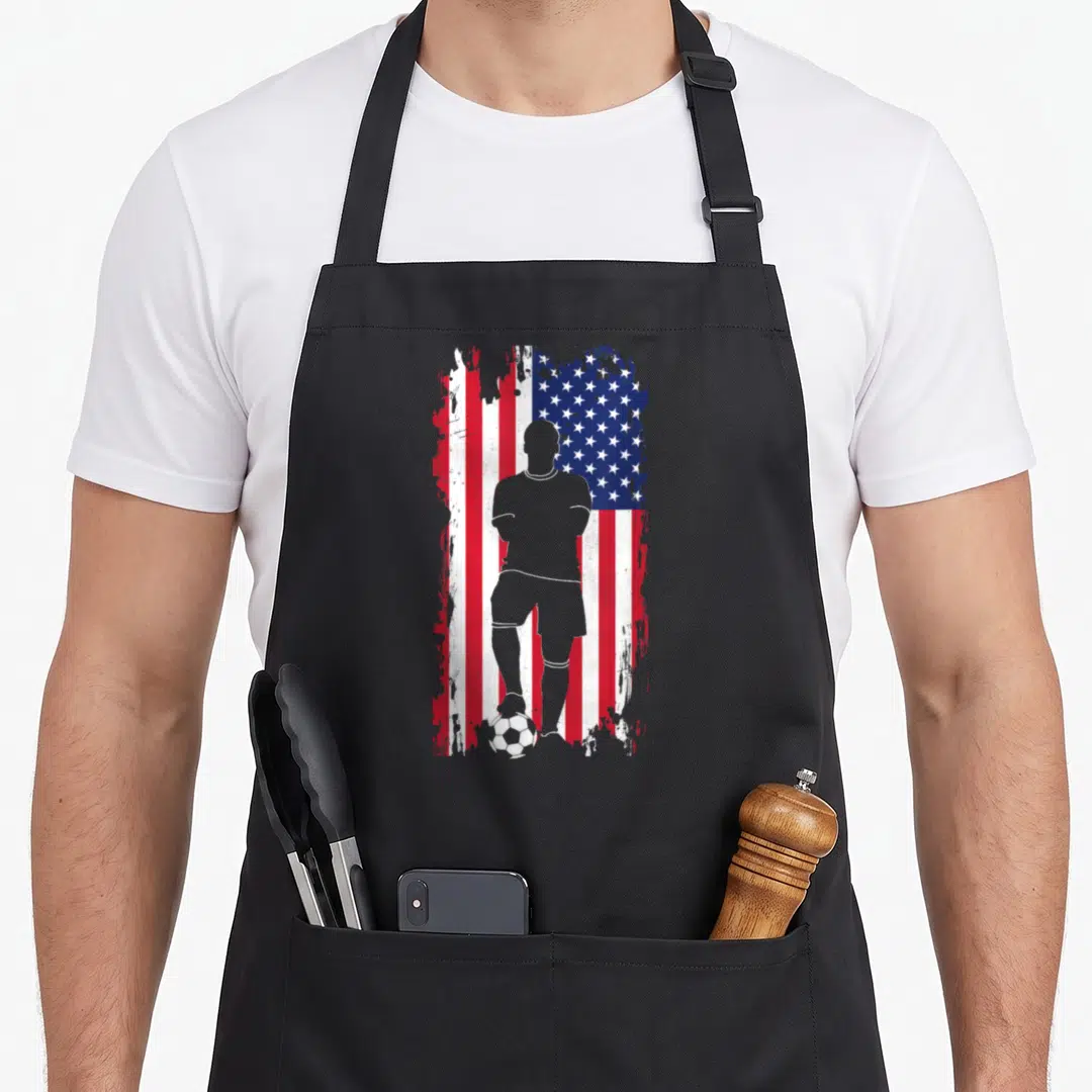 Soccer american flag apron - Grey soccer player silhouette standing on a ball against a distressed red white and blue — shown on a black cotton canvas apron against a white background