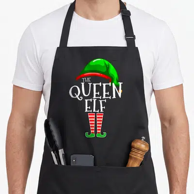 Queen christmas elf apron - White THE QUEEN ELF text between green hat and red striped boots on black background — shown on a black cotton canvas apron against a white background