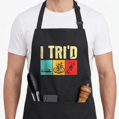 Cyclist triathlete apron - I TRI'D text above teal swimmer, yellow cyclist, and red runner silhouettes — shown on a black cotton canvas apron against a white background