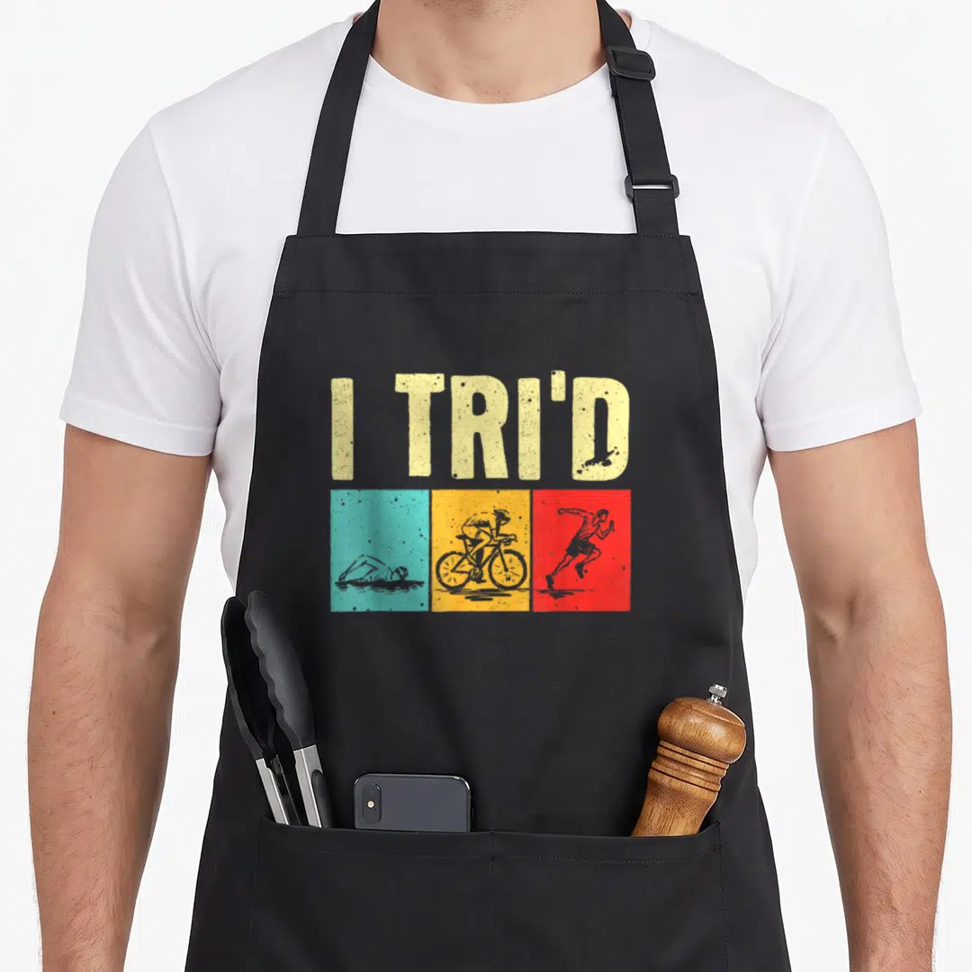 Cyclist triathlete apron - I TRI'D text above teal swimmer, yellow cyclist, and red runner silhouettes — shown on a black cotton canvas apron against a white background