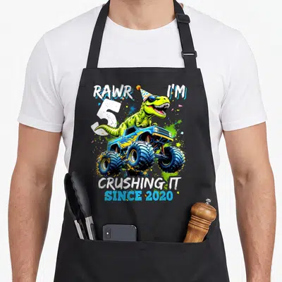 5th birthday monster truck apron - Green dinosaur on blue monster truck with RAWR I'M 5 text — shown on a black cotton canvas apron against a white background
