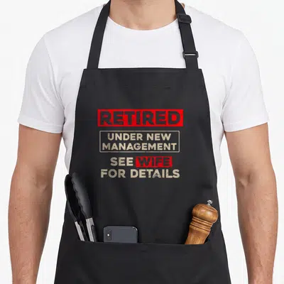 Retirement husband apron - Stacked red and beige text reading Retired Under New Management See Wife For Details — shown on a black cotton canvas apron against a white background