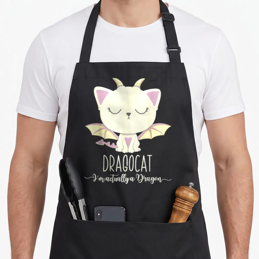 Dragon kitten apron - Cream cat with pink ears and yellow wings above stacked white text reading DRAGOCAT — shown on a black cotton canvas apron against a white background