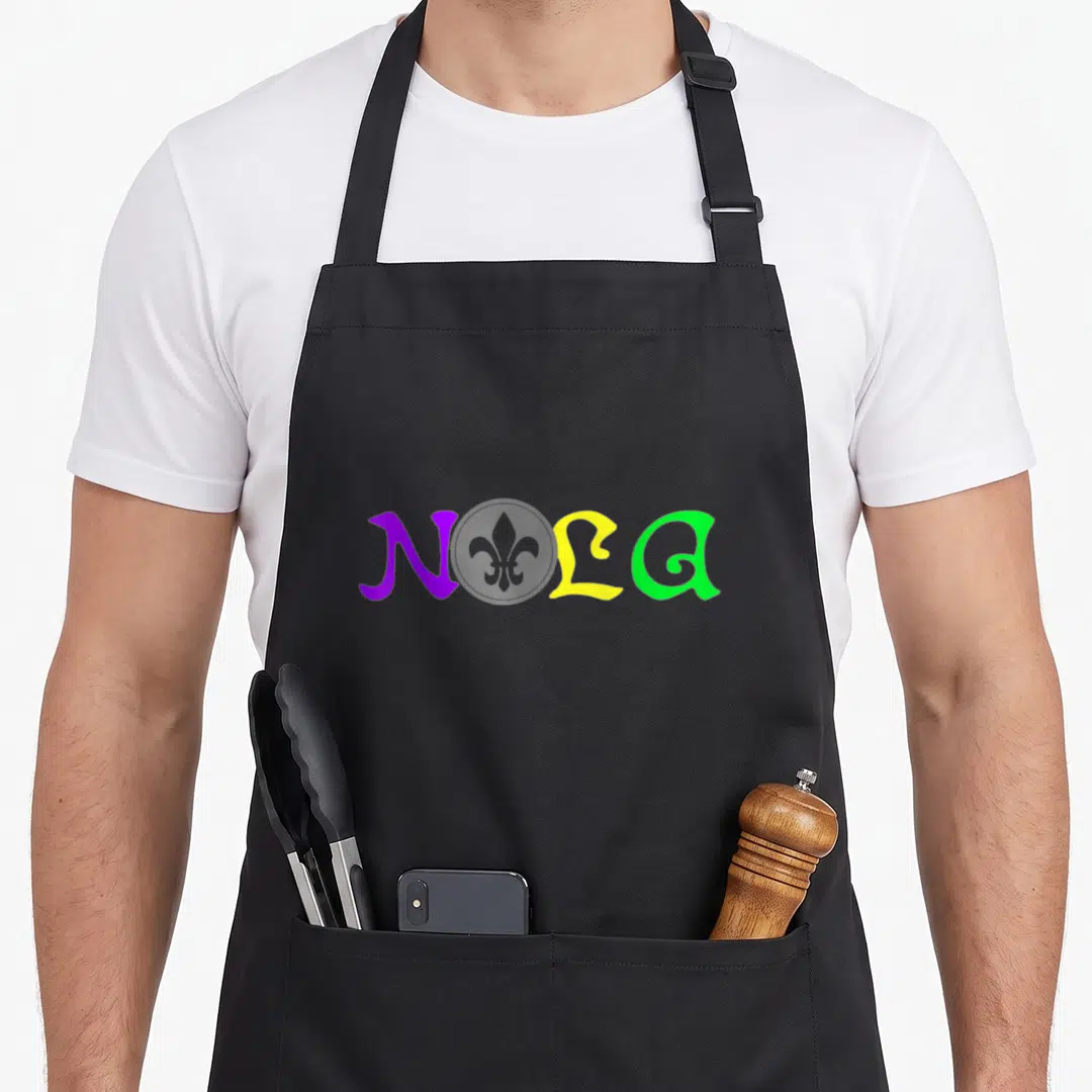 Nola mardi gras aprons - NOLA text in purple yellow green with faint grey fleur de lis behind L — shown on a black cotton canvas apron against a white background