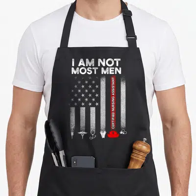Most nursing aprons - I AM NOT MOST MEN text above distressed USA flag with vertical CERTIFIED NURSING ASSISTANT stripe — shown on a black cotton canvas apron against a white background