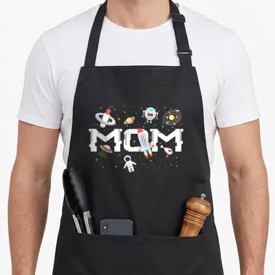 Spaceman birthday apron - MOM text with rocket astronaut robot planets in white red blue yellow cartoon style — shown on a black cotton canvas apron against a white background