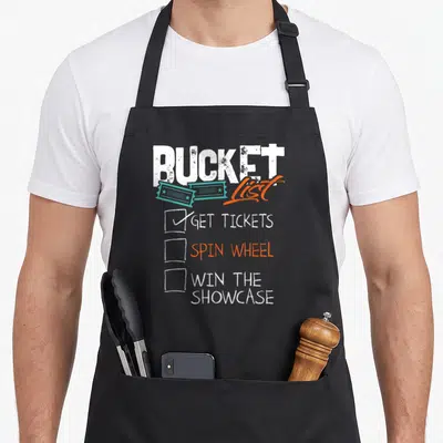Spin bucket apron - BUCKET List text with teal tickets and checklist items GET TICKETS SPIN WHEEL WIN THE SHOWCASE — shown on a black cotton canvas apron against a white background