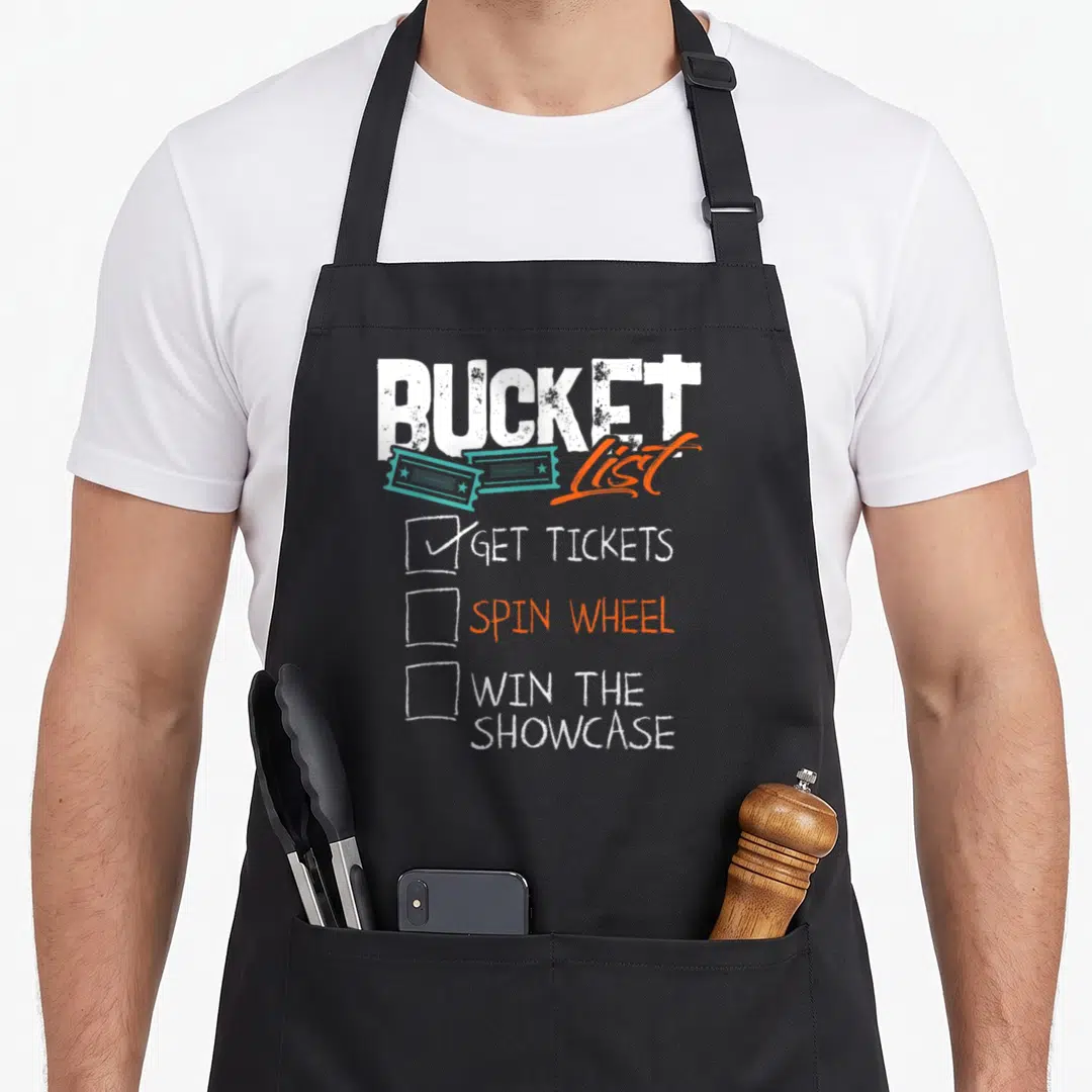 Spin bucket apron - BUCKET List text with teal tickets and checklist items GET TICKETS SPIN WHEEL WIN THE SHOWCASE — shown on a black cotton canvas apron against a white background