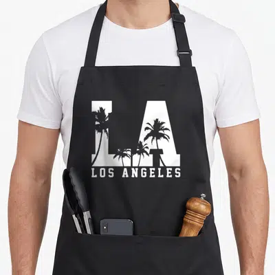 Souvenir los angeles apron - White LA letters with grey palm tree silhouettes and LOS ANGELES text below — shown on a black cotton canvas apron against a white background