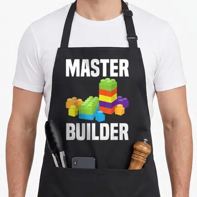 Building grill master apron - MASTER BUILDER text framing cartoon building blocks in green orange yellow purple blue — shown on a black cotton canvas apron against a white background