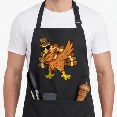 Thanksgiving pilgrim apron - Cartoon turkey wearing pilgrim hat and sunglasses dabbing in brown yellow and black — shown on a black cotton canvas apron against a white background
