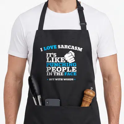 Funny sarcasm face apron - White and blue typography reading I Love Sarcasm with a fist graphic next to IT'S LIKE — shown on a black cotton canvas apron against a white background