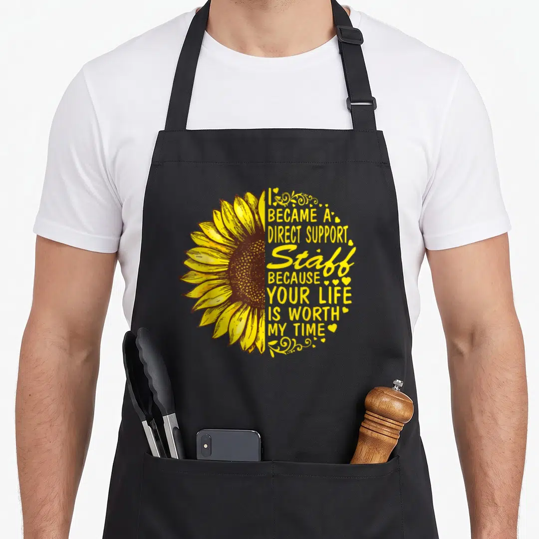 Became sunflower apron - Half sunflower with stacked yellow text and hearts forming a circle — shown on a black cotton canvas apron against a white background